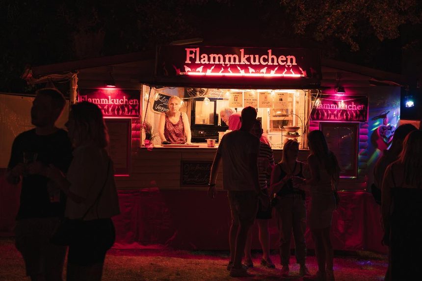 Beleuchteter Flammkuchenstand am Abend, davor wartende Gäste